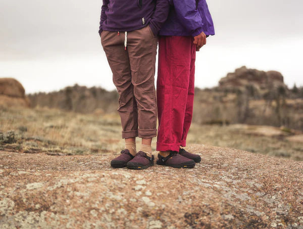 Two people stand back-to-back outdoors on rocks, each wearing Bedrock's Mountain Clog shoes paired with purple tops and loose pants. An overcast sky and blurred rocks form the background.