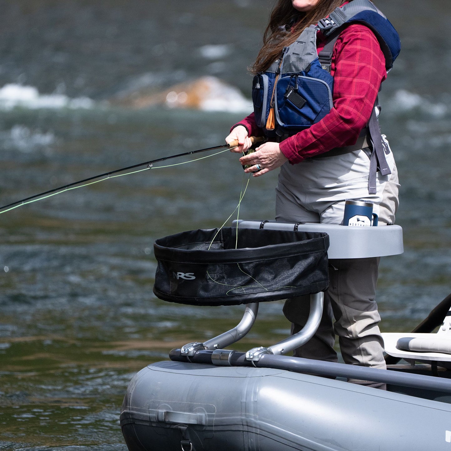 A person wearing a life jacket and holding a fishing rod stands on an inflatable raft in a river, preparing to fish, equipped with the NRS Stripping Basket for Molded Thigh Hooks.