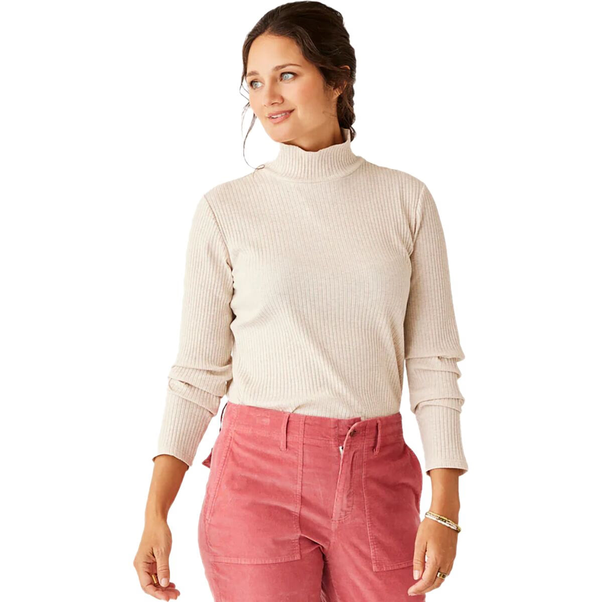 A woman stands against a plain background, wearing the Carve Denise Turtleneck in light beige ribbed fabric paired with high-waisted pink corduroy pants, looking to the side.