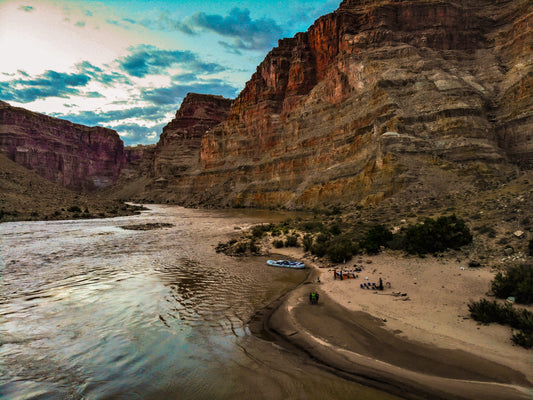 A group enjoys dusk on a sandy riverbank in Cataract Canyon after their Intro to Big Water Kayaking trip with 4CRS Paddle School, with kayaks docked nearby and steep canyon walls rising above.