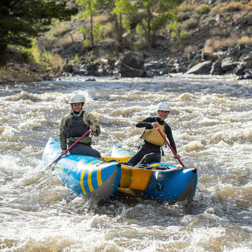 Two people wearing helmets and life jackets paddle the blue Badfish Hammer Cat inflatable boat, made of DuraClear fabric, through whitewater rapids on a river lined with rocks and trees.