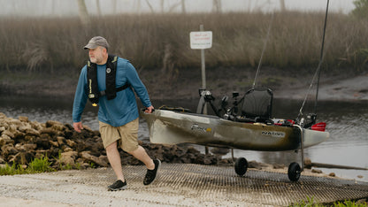 A man wearing a cap and life jacket effortlessly transports his Native Watercraft kayak, equipped with fishing rods, using the SideKick Kayak Wheel System at a boat ramp near a foggy marsh.
