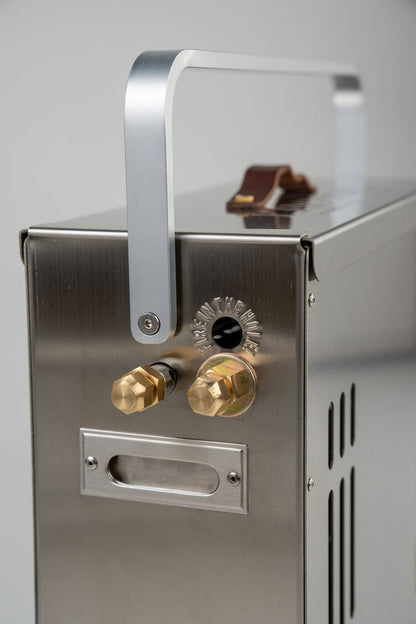 Close-up of the LavaBox BoomBox Portable Campfire, featuring a stainless steel body, metal handle, two brass fittings, and side ventilation slots.
