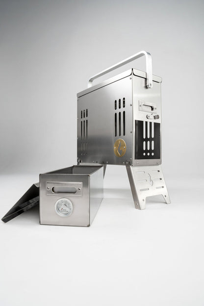 The LavaBox BoomBox Portable Campfire is a stainless steel grill with a handle and metal box featuring a built-in thermometer, offering dual-flame, smokeless campfires anywhere.