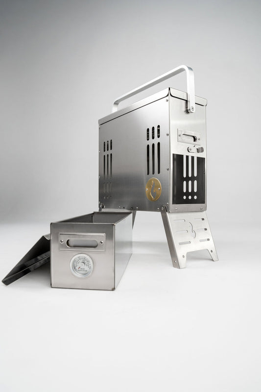 The LavaBox BoomBox Portable Campfire is a stainless steel grill with a handle and metal box featuring a built-in thermometer, offering dual-flame, smokeless campfires anywhere.