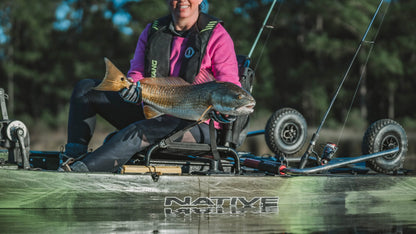 A person in a kayak holds a large fish while wearing a pink jacket and life vest, surrounded by fishing gear. Thanks to the Native Watercraft SideKick Kayak Wheel System, their kayak is effortlessly transported to this prime fishing spot.