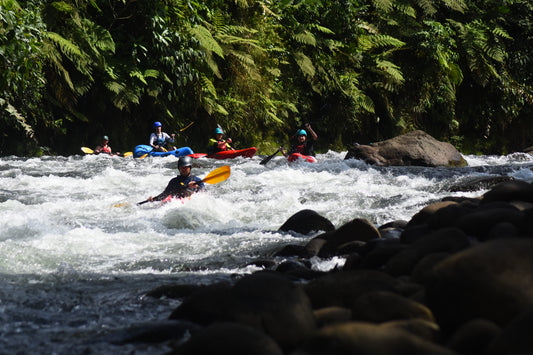Five kayakers from 4CRS Paddle School navigate through rapids in a river surrounded by lush green vegetation and rocks during the Costa Rica Paddling Trip.