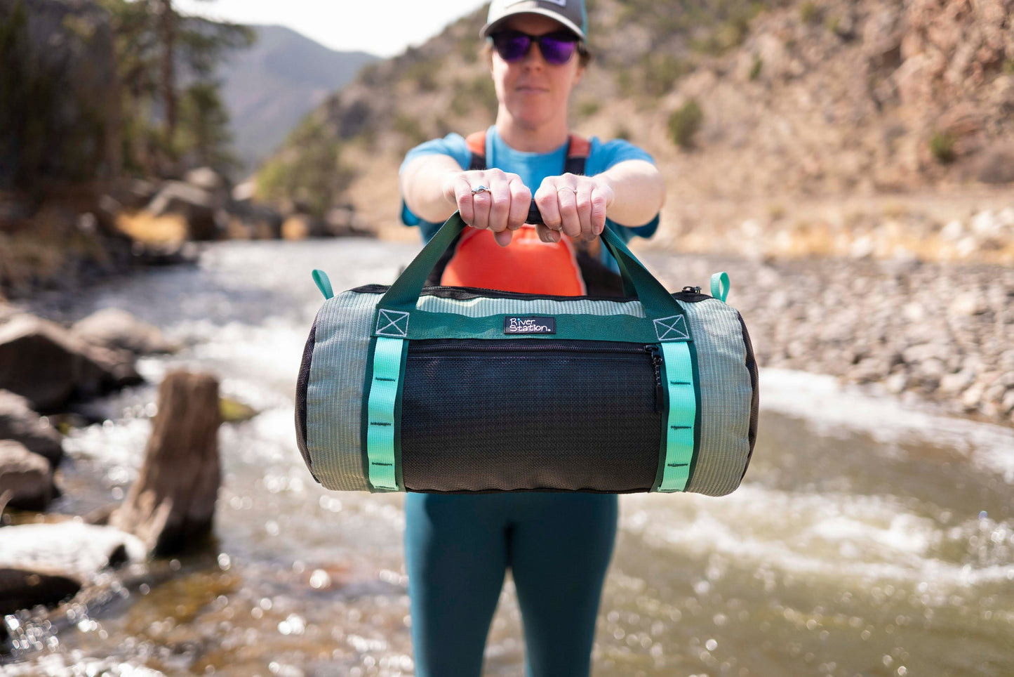 A person stands by a rocky river holding a River Station Gear Small Mesh Duffel with both hands extended toward the camera.