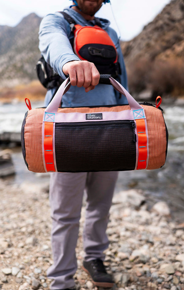 A person stands on a rocky riverbank holding the River Station Gear Small Mesh Duffel, an American-made whitewater bag with orange and black panels and white handles. Mountains and water provide a scenic backdrop.