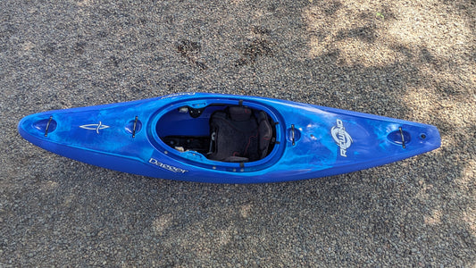 A blue Demo Rewind LG by Dagger, used as a demo kayak, rests on gravel in this overhead view. It features a spacious cockpit and visible footrests, offering comfort and performance for paddlers.