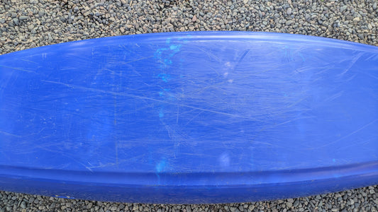 A scratched blue surfboard rests on small, round pebbles beside a used Dagger Demo Rewind LG kayak.