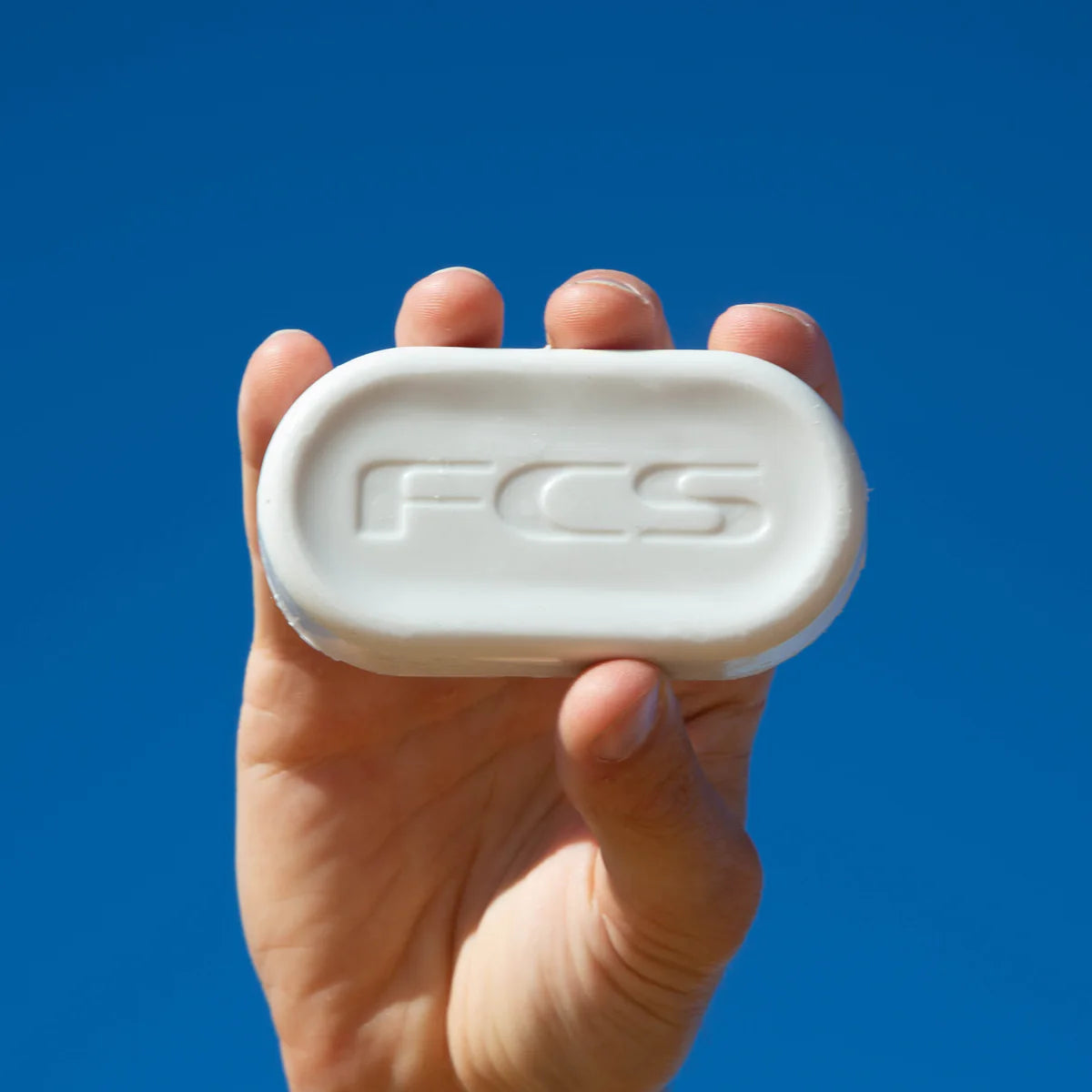 A person holding up a white Surfboard Wax with the word FCS on it.