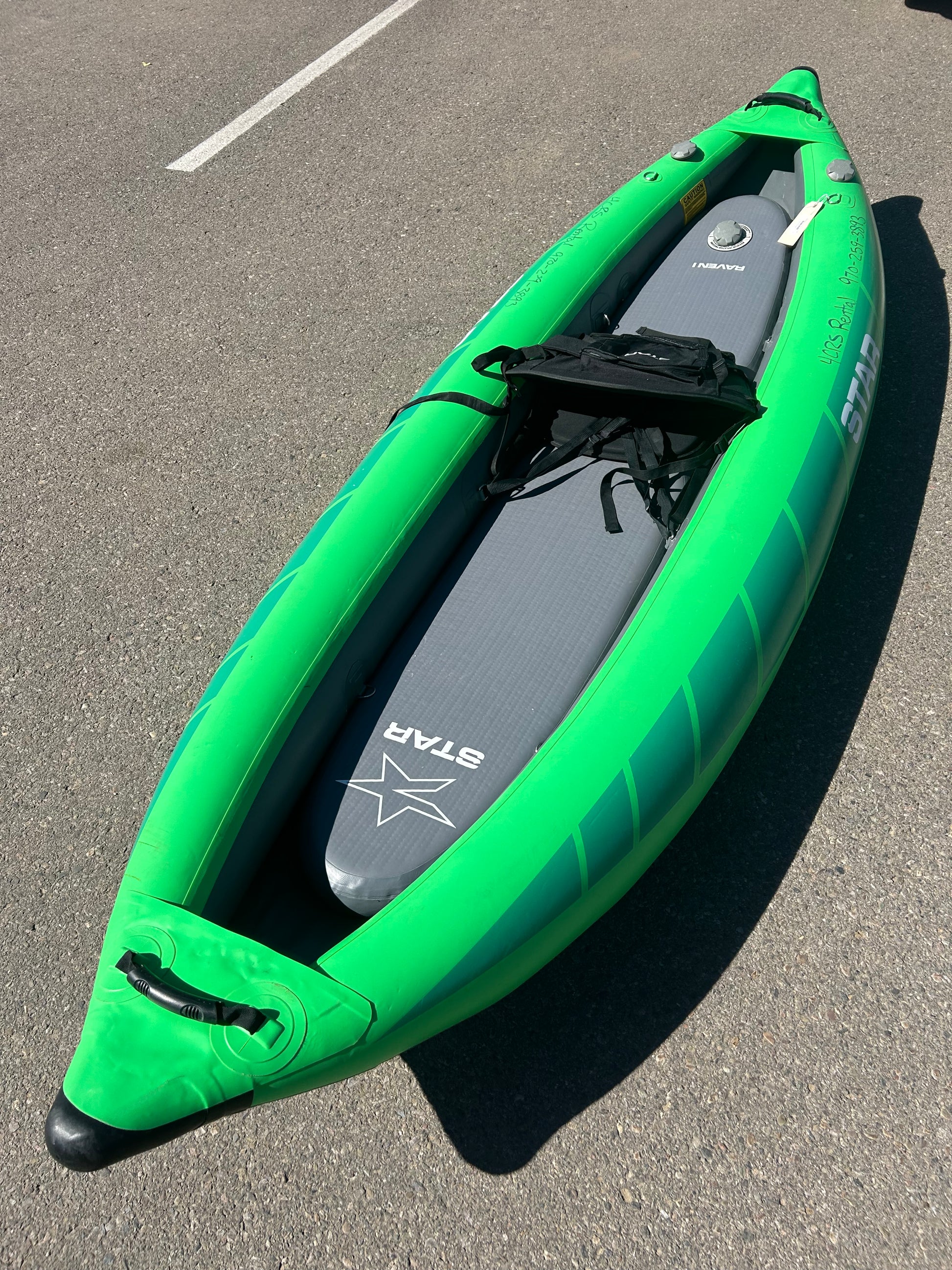 A green inflatable kayak, a used Demo STAR Raven Solo by NRS, is parked on an asphalt surface with a grey paddleboard labeled "STAR" stored inside. This rental demo boat also comes with a convenient carry bag.