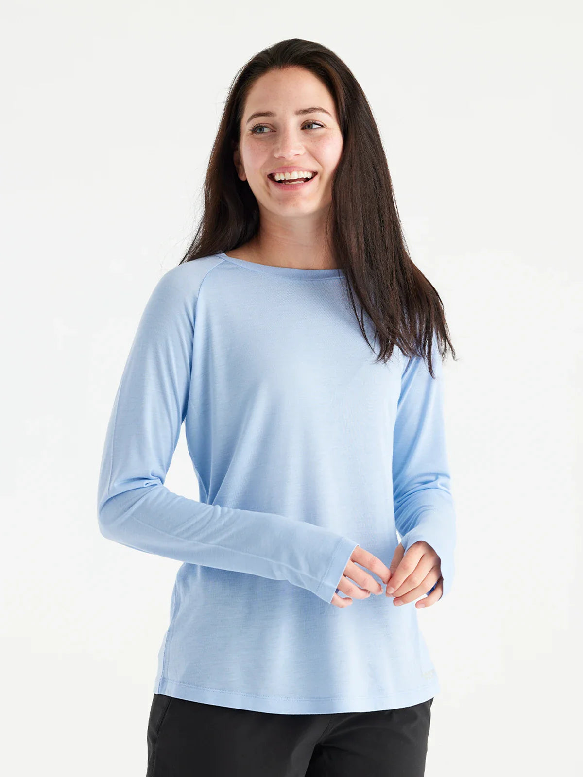 A woman with long dark hair wears the Free Fly Bamboo Lightweight Long Sleeve W's shirt and black pants, standing and smiling against a plain white background.