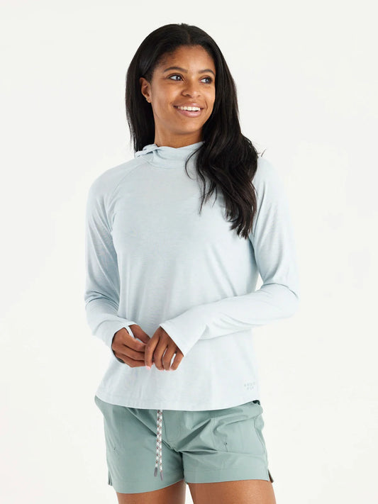 A woman wears the Free Fly Elevate Hoodie W's in light blue with drirelease tech and green shorts, standing against a plain white background, smiling and looking off to the side.