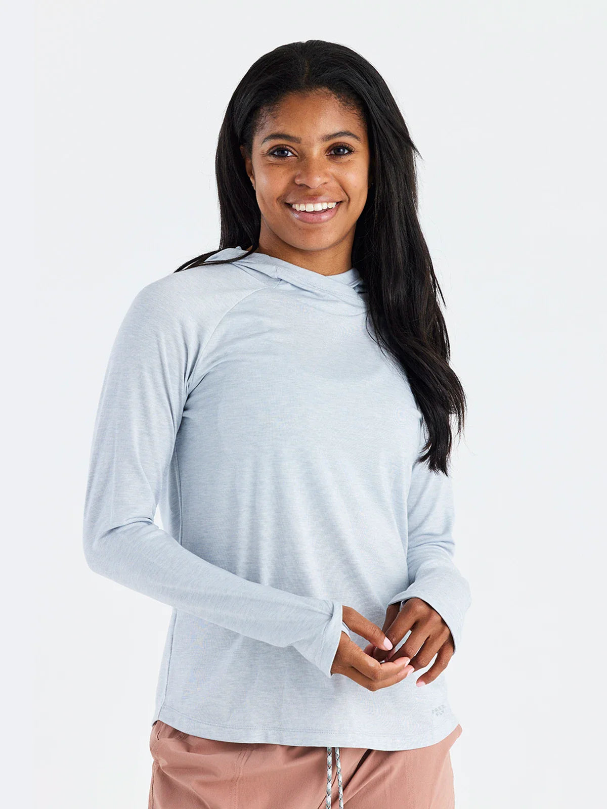 A woman with long dark hair smiles while wearing a light gray Free Fly Elevate Hoodie W's featuring drirelease tech and pink pants, standing against a plain white background.