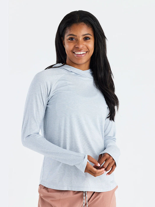 A woman with long dark hair smiles while wearing a light gray Free Fly Elevate Hoodie W's featuring drirelease tech and pink pants, standing against a plain white background.