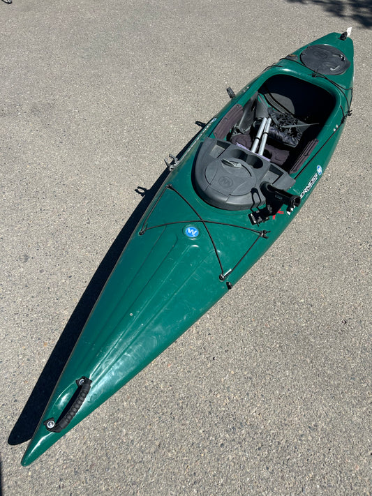 A pre-owned Consignment Pungo 120 green kayak from Wilderness Systems, complete with a paddle, lies on a concrete surface. This kayak boasts an open cockpit, gear storage areas, and a convenient rod holder.