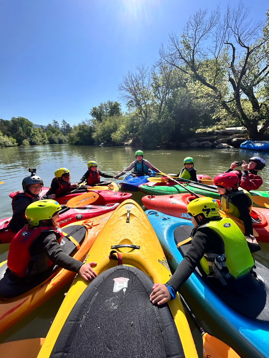 Kids wearing helmets and life jackets sit in colorful kayaks, forming a circle on a calm river, enjoying the Paddle and Paint Adventure Camp by 4CRS Paddle School under a clear blue sky.