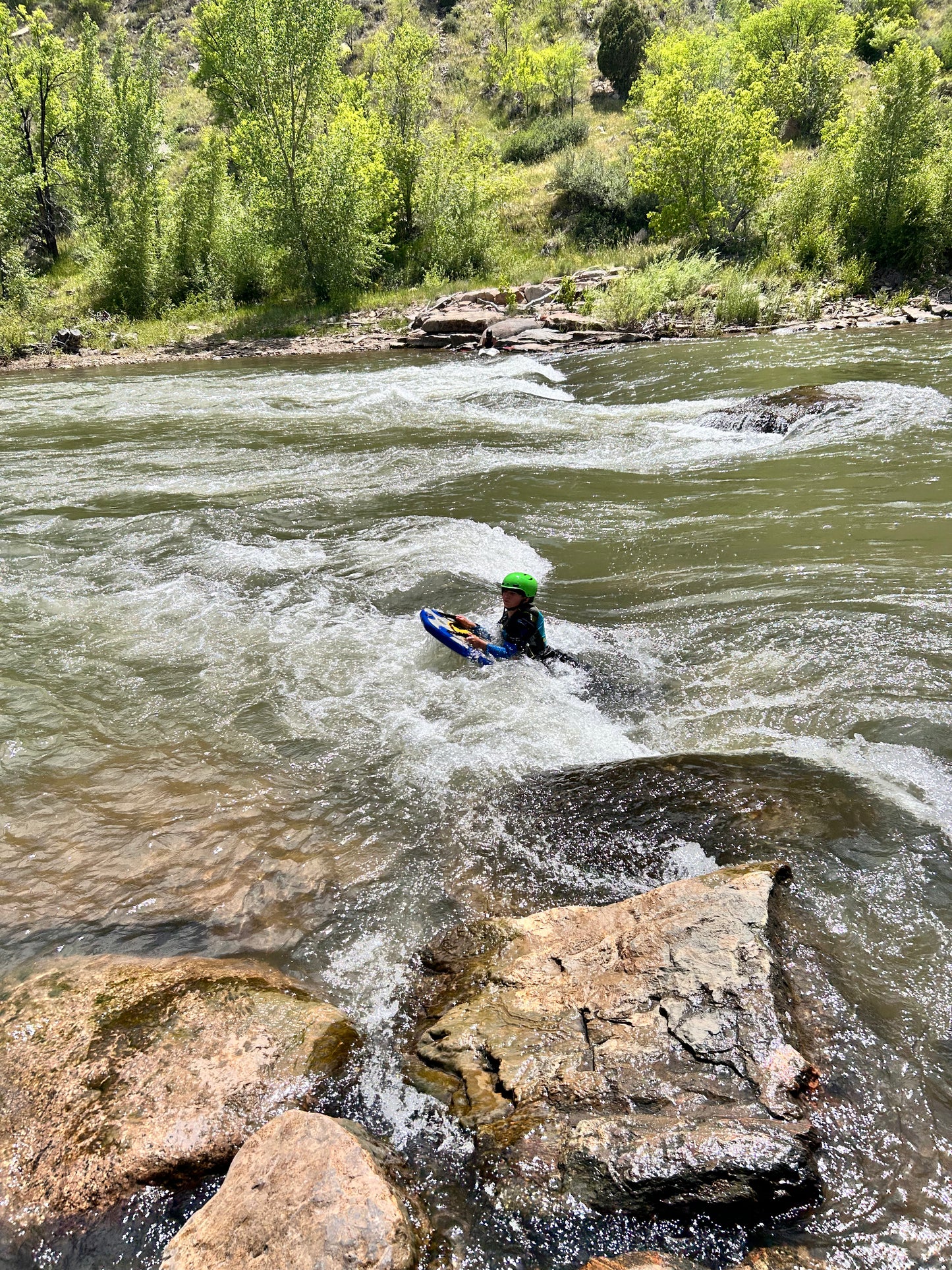 Wearing a green helmet, a kayaker grips a bodyboard and navigates rocky river rapids, capturing the thrill of adventure in 4CRS Paddle School’s Send-It Summer Finale.