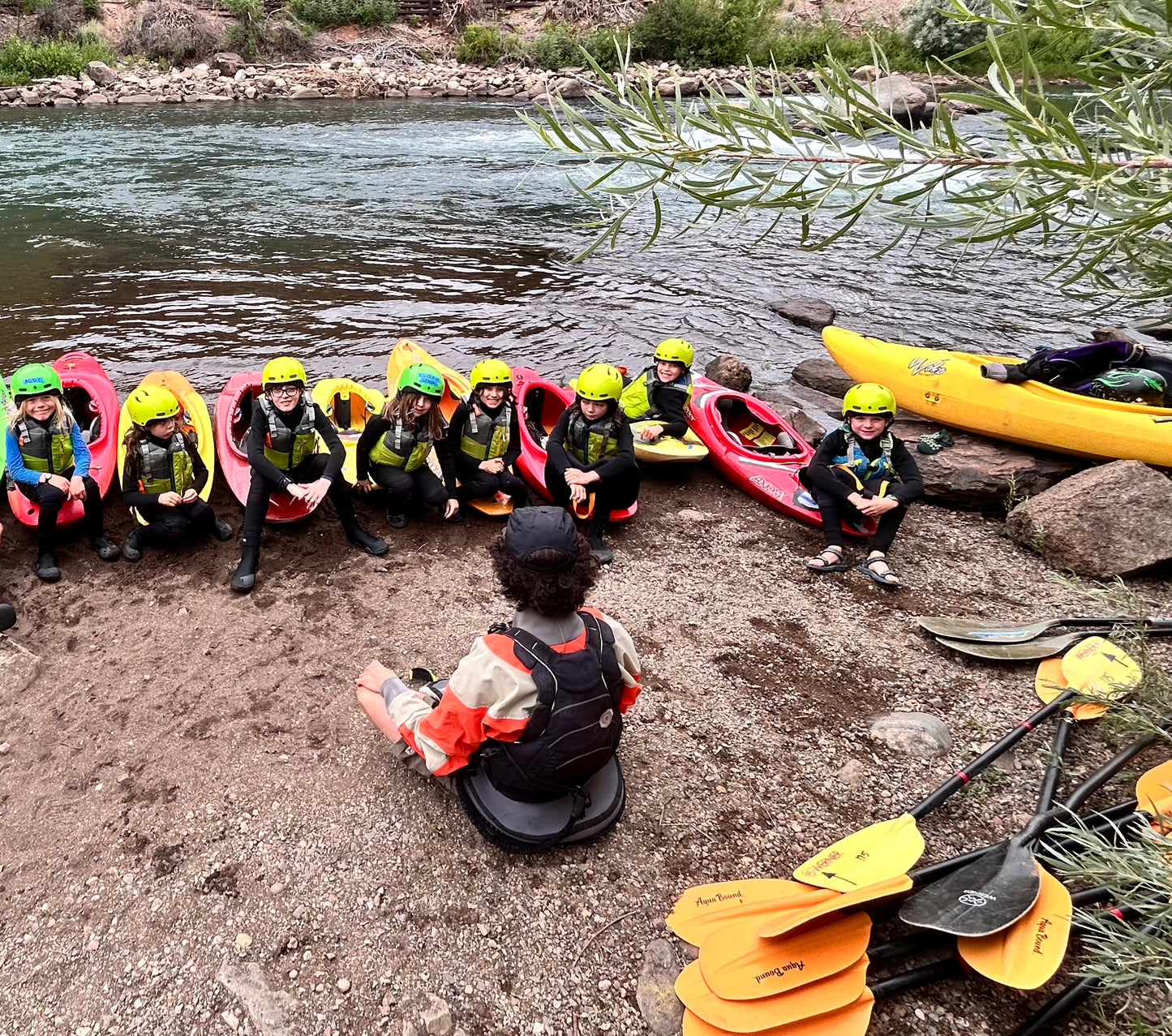 Participants in "Roots to Rapids: Science on the River" by 4CRS Paddle School sit in kayaks on a riverbank, wearing helmets and wetsuits, as an instructor teaches river safety. Yellow paddles and extra kayaks are nearby.