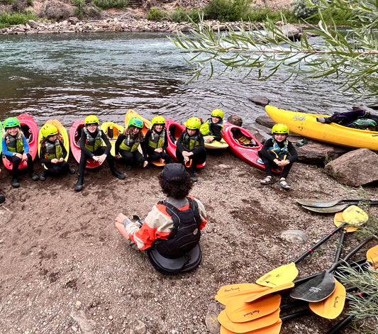 Participants in "Roots to Rapids: Science on the River" by 4CRS Paddle School sit in kayaks on a riverbank, wearing helmets and wetsuits, as an instructor teaches river safety. Yellow paddles and extra kayaks are nearby.