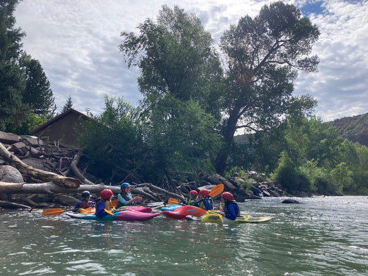 A class of kids from the 4CRS Paddle School paddling in a river near a cabin with their Little Kids Level 2 Kayak Class.