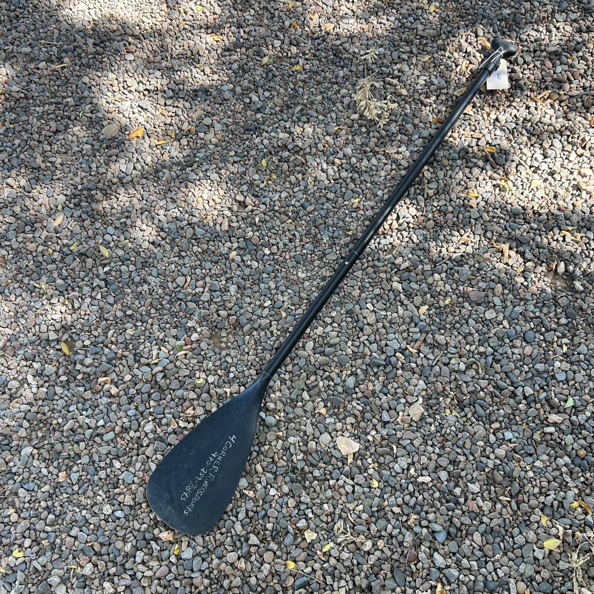The Badfish Demo SUP Paddle 70-85, featuring a wide blade, rests on gravel in an outdoor setting.