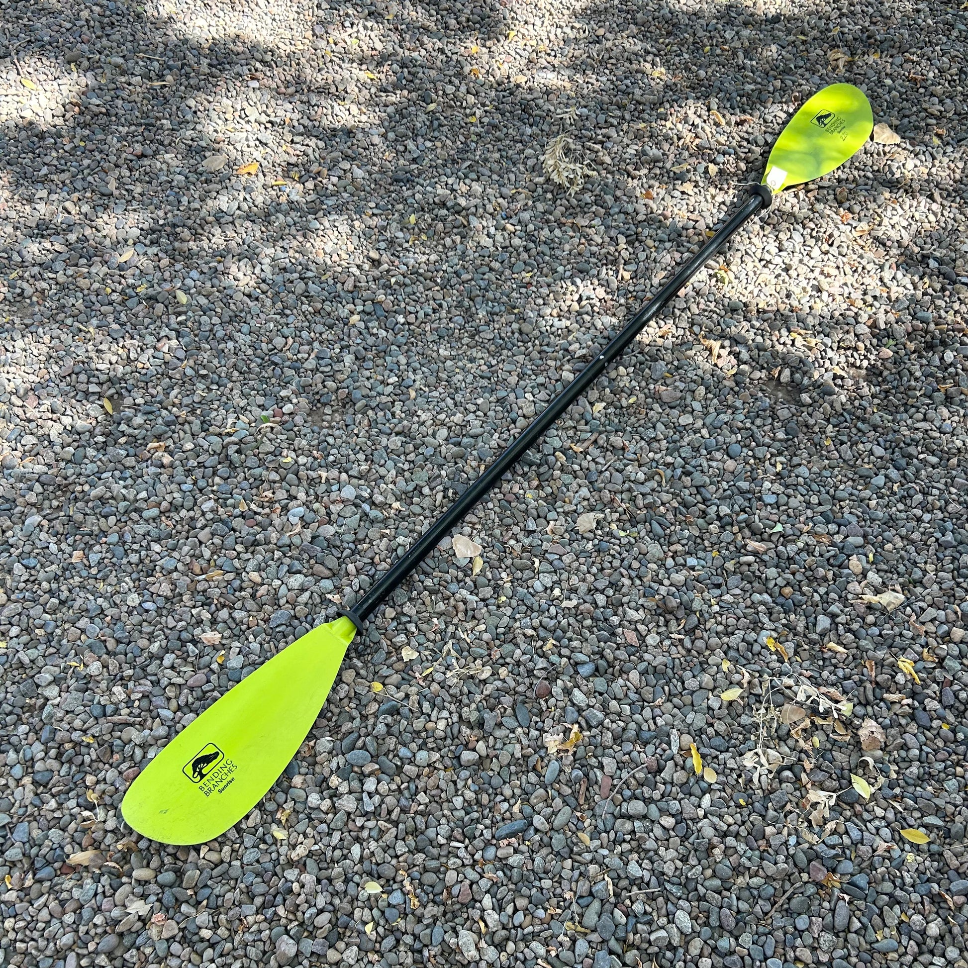 The AquaBound Demo Sunrise fiberglass kayak paddle, featuring yellow blades and a black shaft, lies on gravel ground, ideal for flatwater kayaking or recreational paddling.