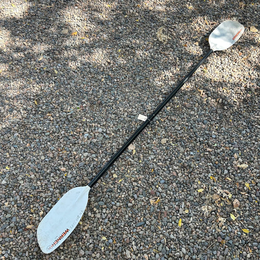 A Werner Demo Rio 2 Piece Whitewater Kayak Paddle, equipped with river running blades and a robust fiberglass shaft with white blades, lies on a gravel surface.