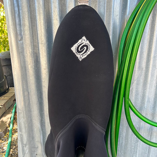 A black Snap Dragon Demo River Trek Skirt with a visible brand patch hangs on a corrugated metal wall beside coiled green garden hoses, ready for the next River Trek.