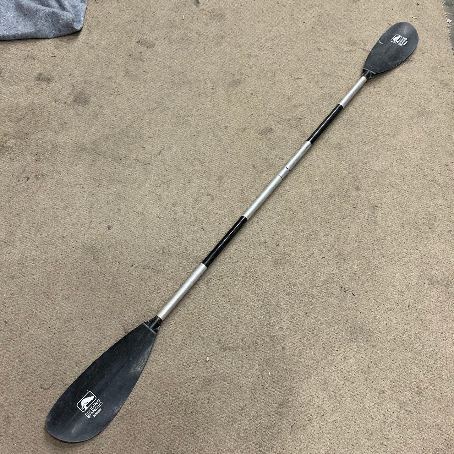 A black and silver Demo Whisper Paddle by AquaBound with two blades lies on a beige carpeted surface. This entry-level kayak paddle features adjustable feathering angles and a durable aluminum shaft, making it perfect for novice paddlers.