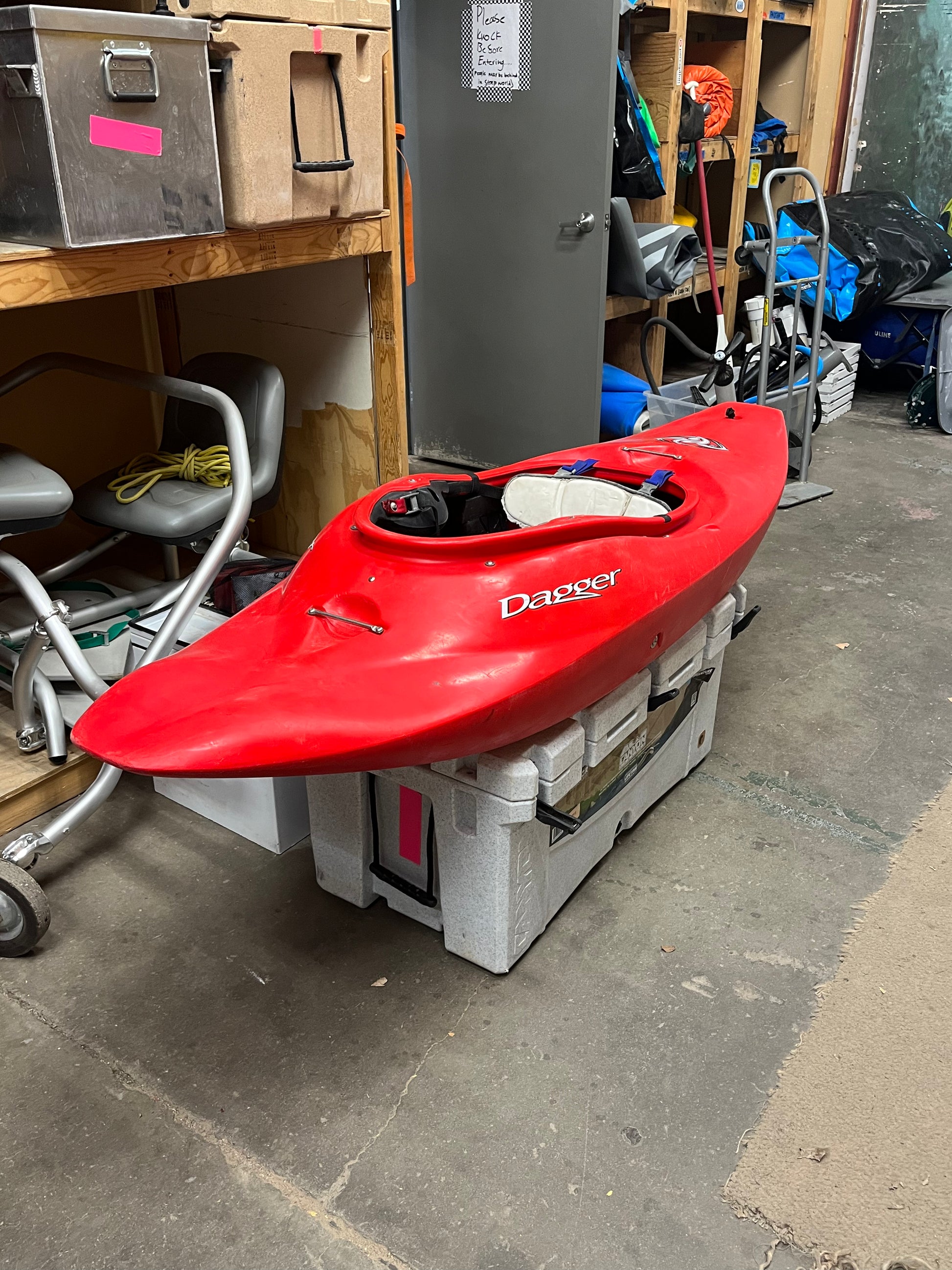 A Consignment Dagger ID 7.0 kayak, in red, is carefully balanced atop large plastic boxes in a cluttered storage room, showcasing its well-maintained condition.
