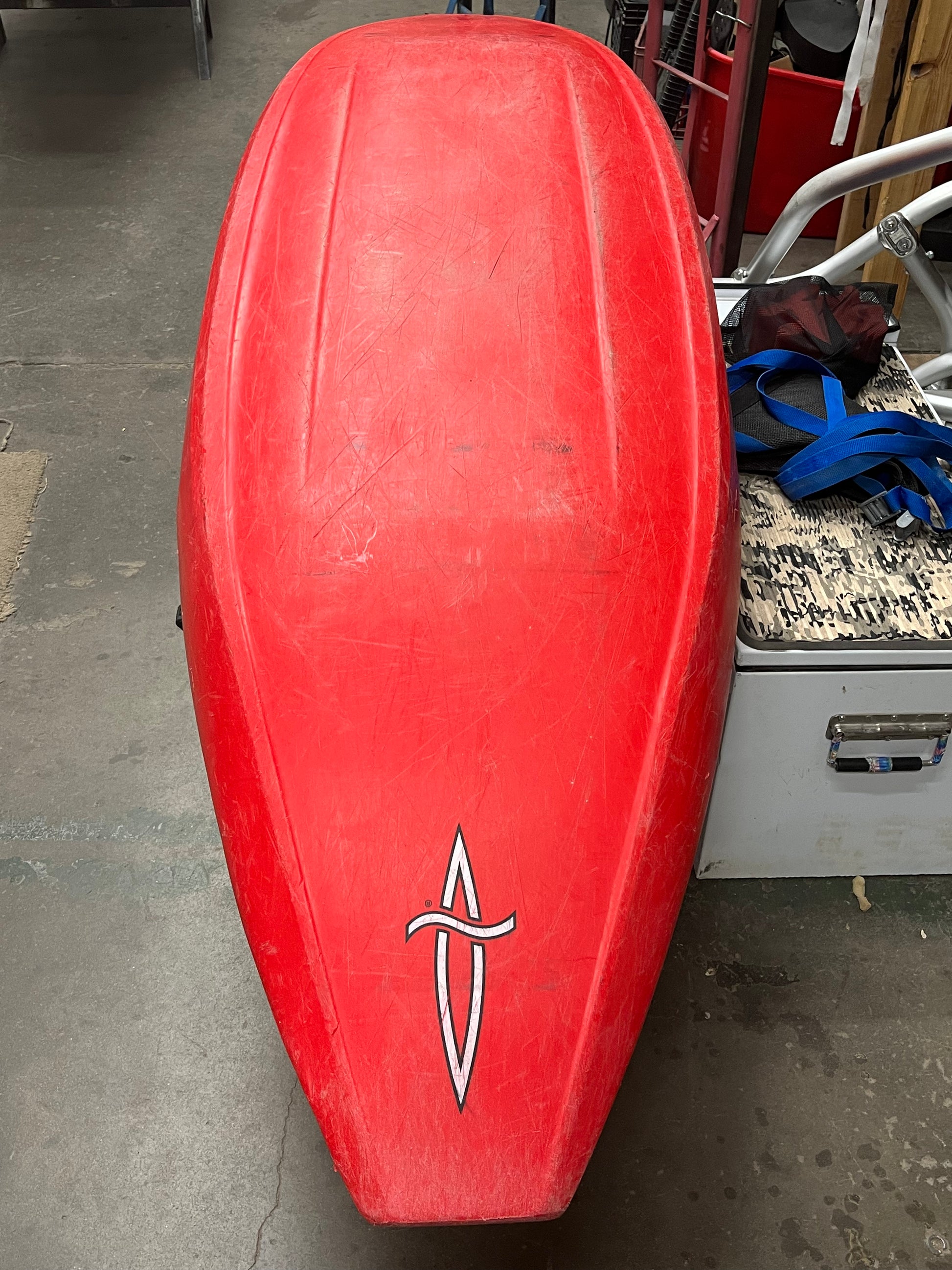 A Consignment Dagger ID 7.0 kayak lies upside down, its bottom side showing a logo near the stern. Blue straps and a storage box are nearby, indicating the kayak's ready-for-adventure state with no concerns for shipping costs.