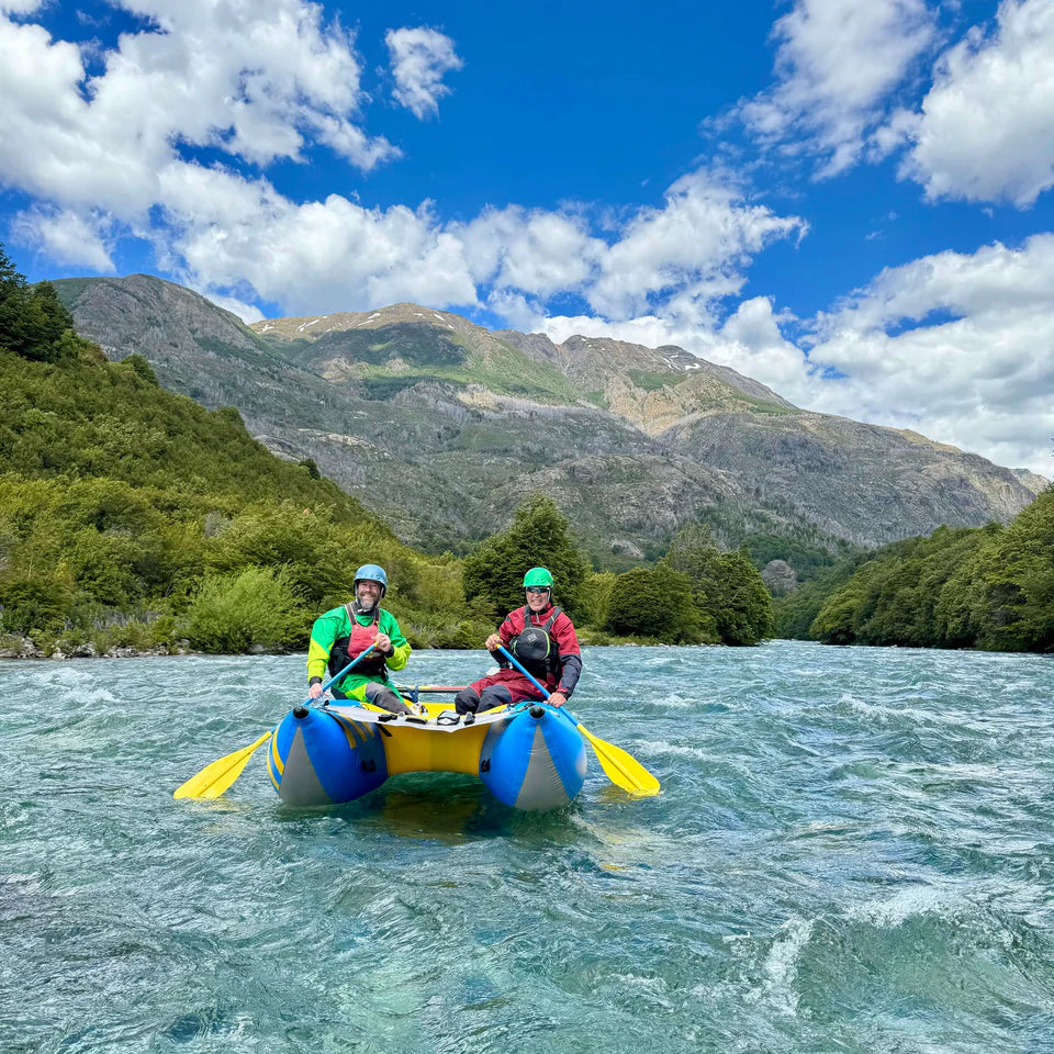 Two people in helmets and life jackets paddle a yellow Badfish Hammer Cat inflatable boat on a river, with green hills and mountains in the background under a partly cloudy sky.