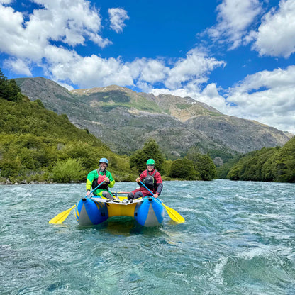 Two people in helmets and life jackets paddle a yellow Badfish Hammer Cat inflatable boat on a river, with green hills and mountains in the background under a partly cloudy sky.