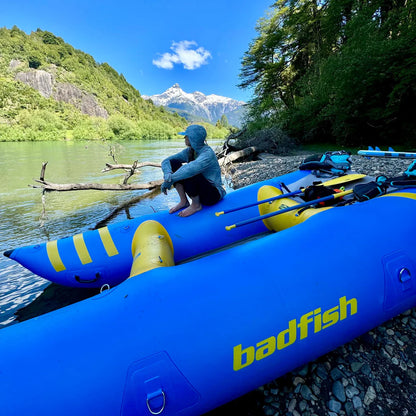 A person sits on a riverbank beside a bright blue Badfish Hammer Cat inflatable boat, with paddles by their side and snow-capped mountains in the background.
