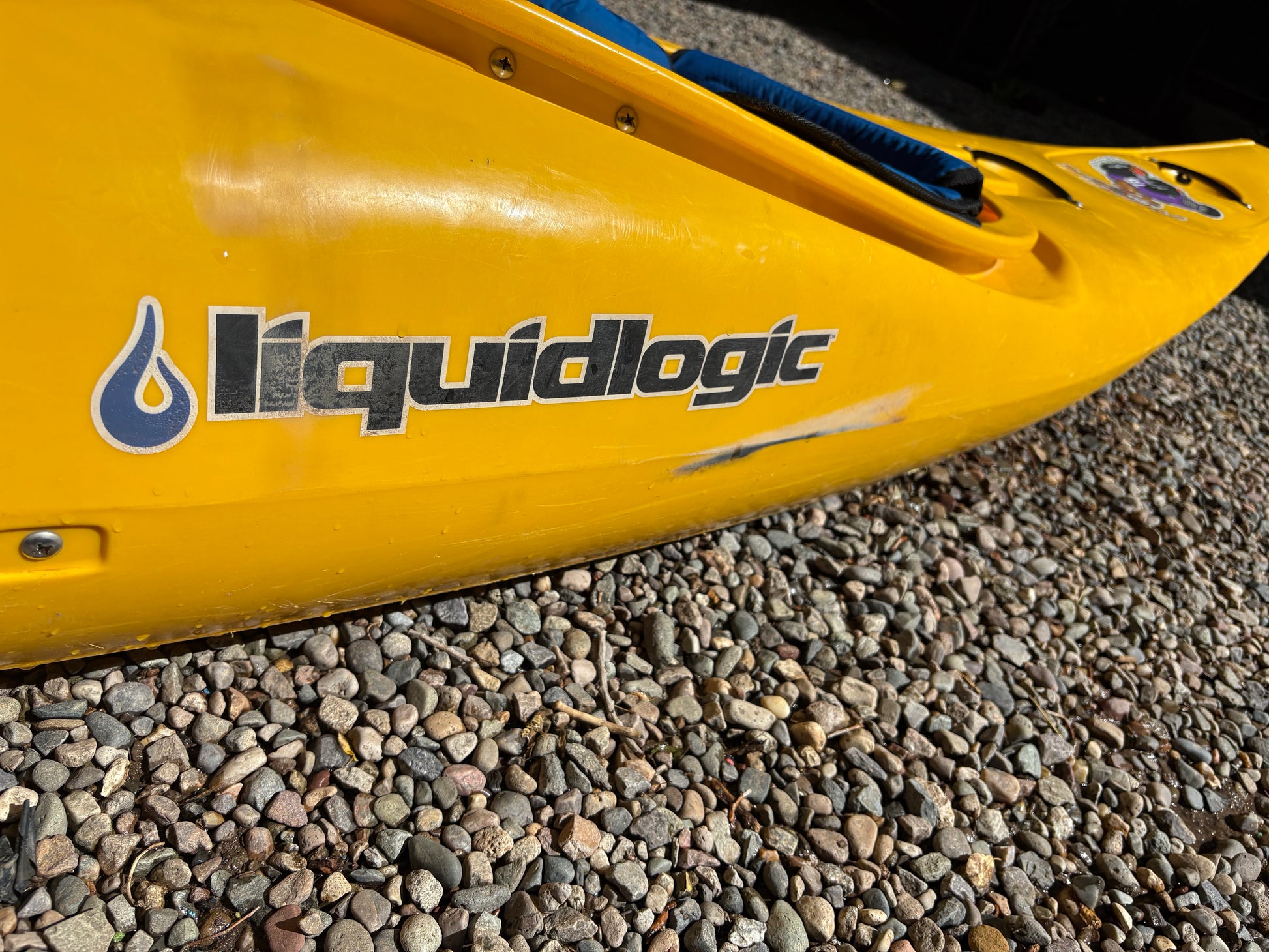 Close-up of the yellow LiquidLogic Demo Powerslide kayak with logo, resting on pebbles—ideal as a demo boat or for anyone seeking a quality used kayak.