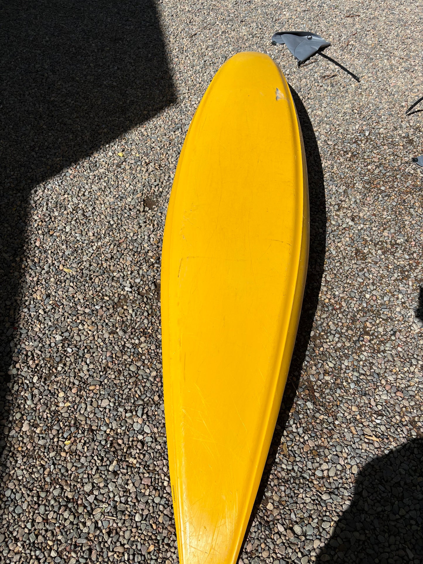 A yellow surfboard with a detached black fin lies on gravel beside a used kayak and a LiquidLogic Demo Powerslide boat.