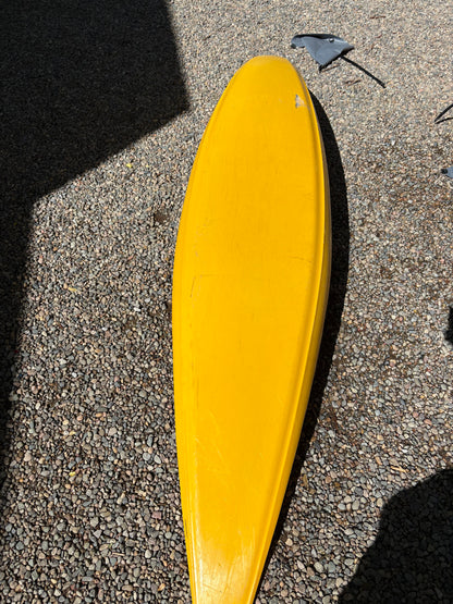 A yellow surfboard with a detached black fin lies on gravel beside a used kayak and a LiquidLogic Demo Powerslide boat.