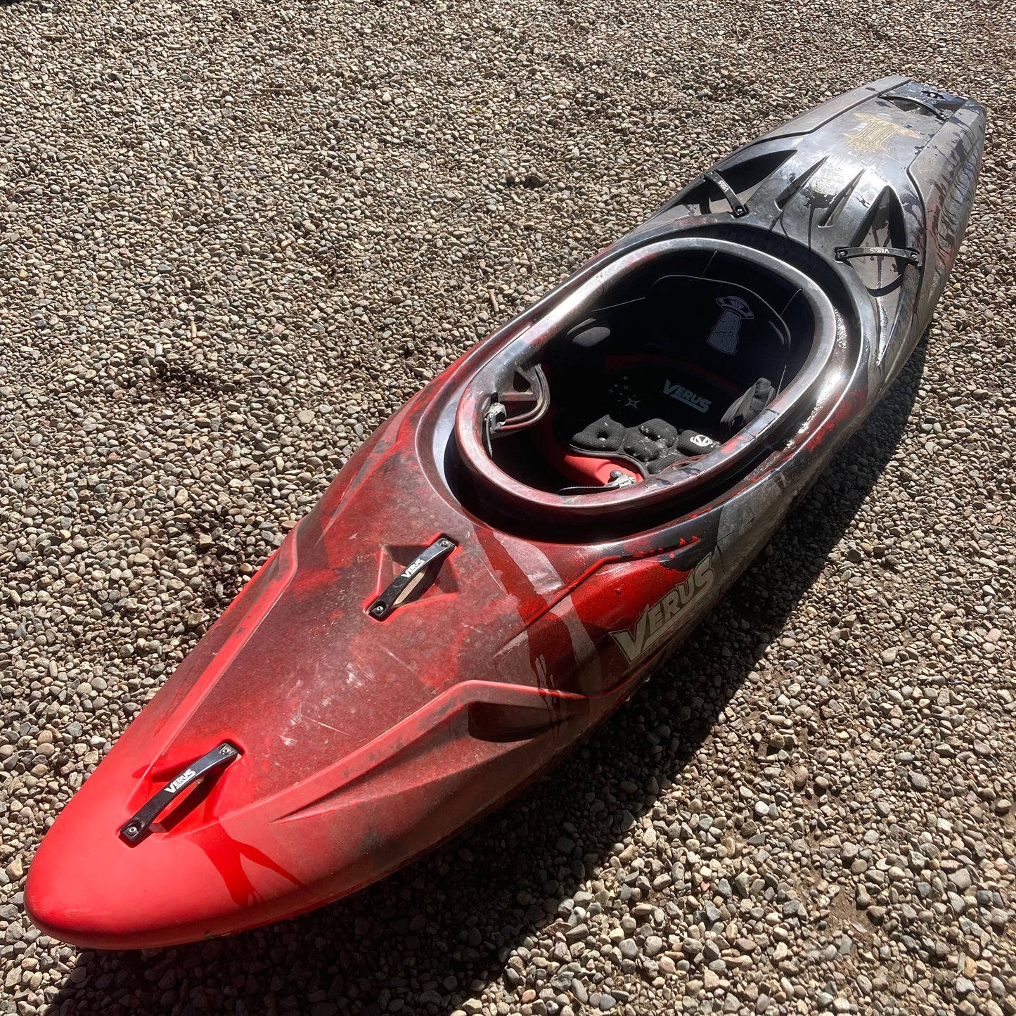 A red and black Demo Flux M/L kayak by Verus laying on gravel.