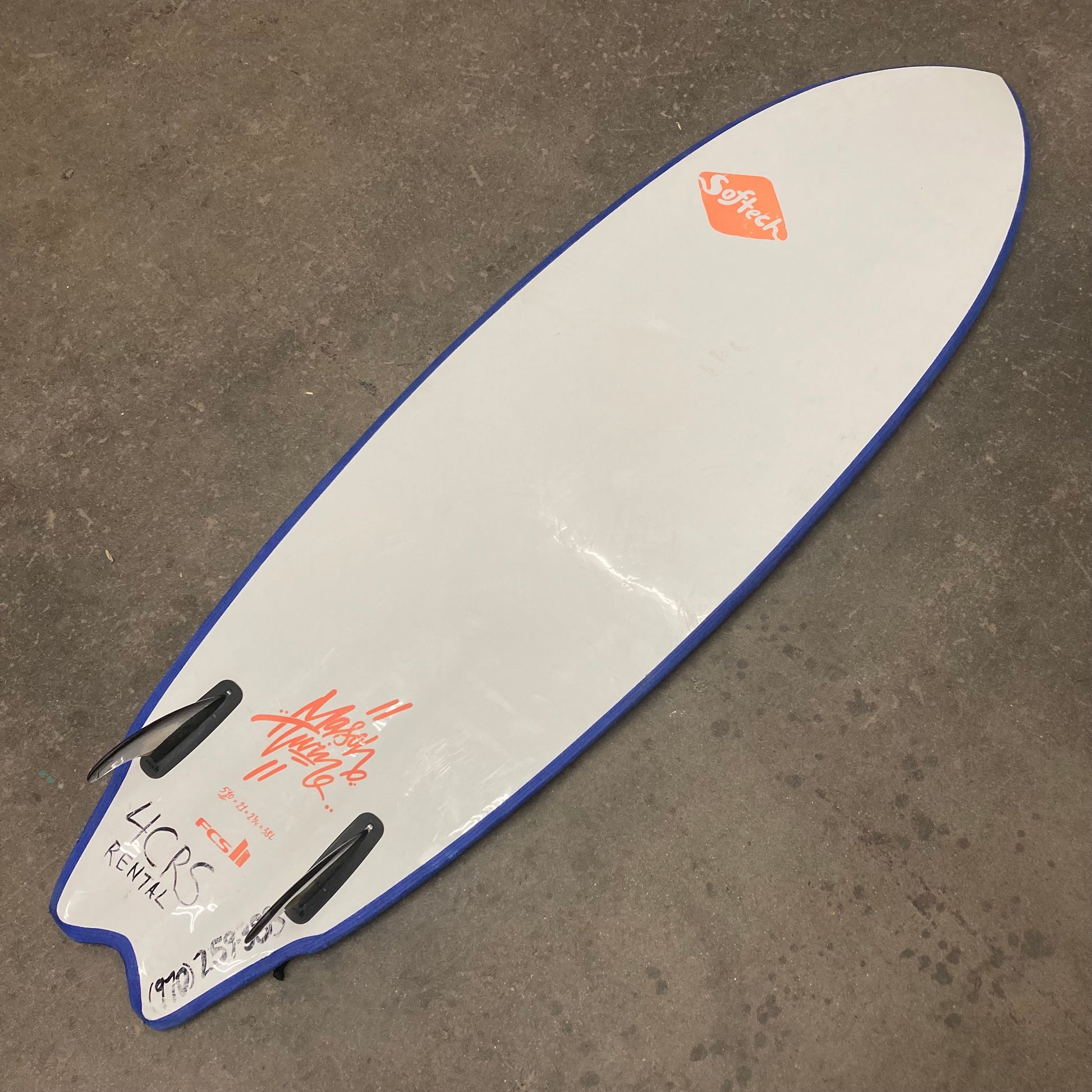 A white and blue Demo Mason Twin surfboard laying on a concrete floor. (Brand: FCS)