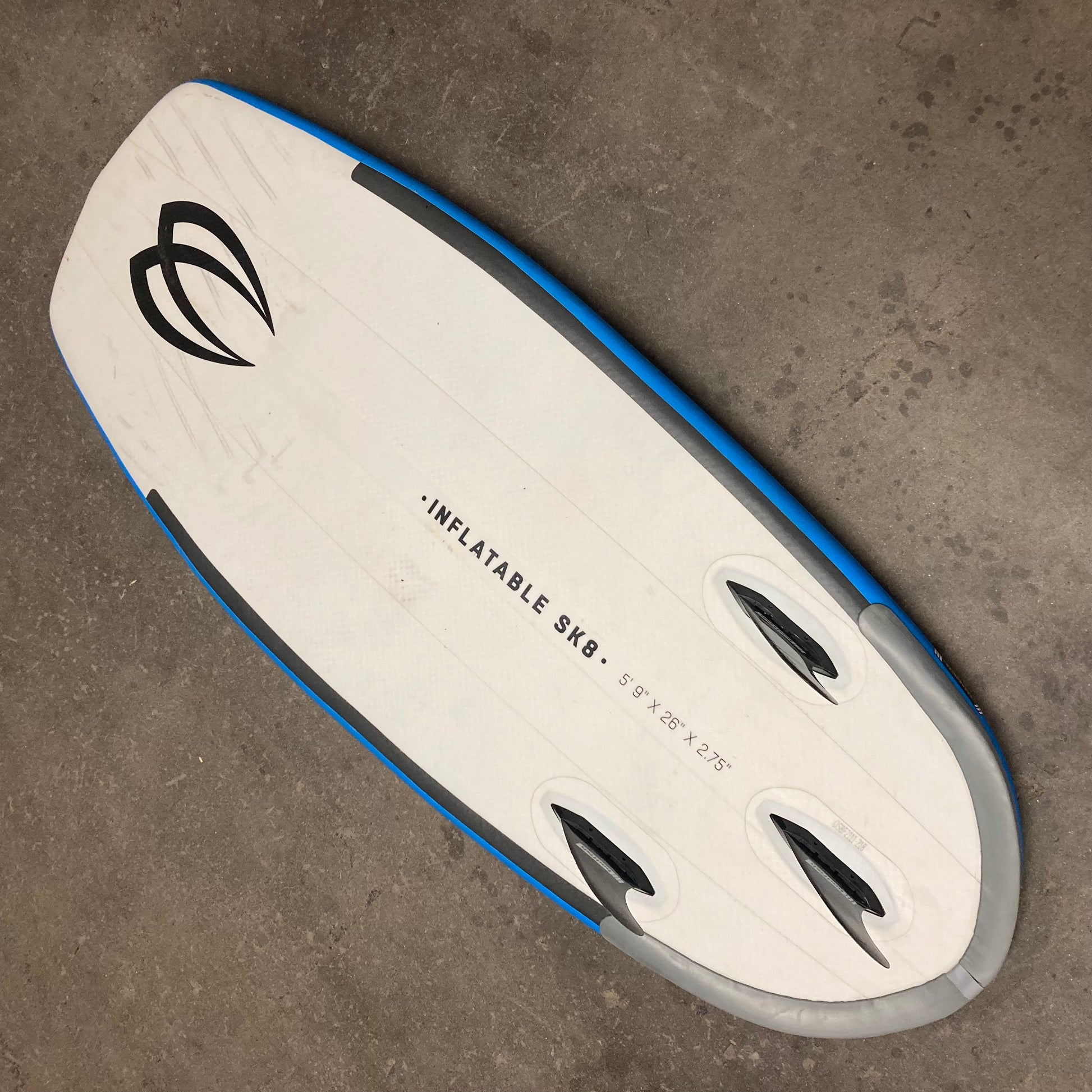 A white and blue Demo ISK8 surfboard laying on a concrete floor. (Brand: Badfish)