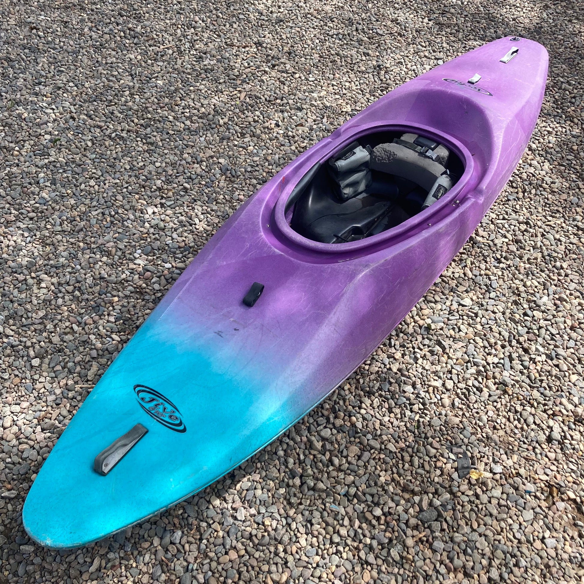 A Consignment Necky Jive kayak in purple and blue laying on gravel.