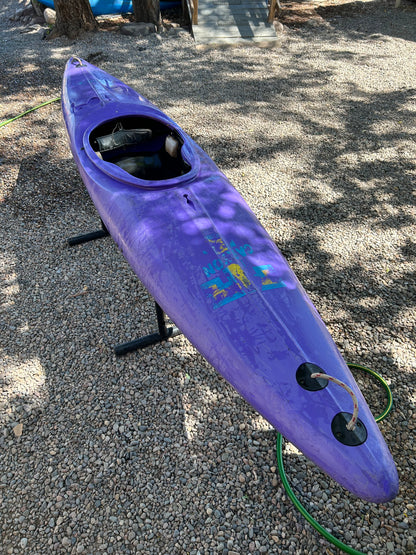 A Used Prijon Canyon kayak by 4Corners Riversports, crafted from durable HTP plastic in purple, sits on a stand over gravel with a garden hose nearby and tree shadows on the ground.
