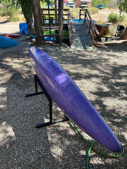 A Used Prijon Canyon kayak from 4Corners Riversports, crafted with durable HTP plastic, sits on a metal stand over gravel with a hose beside it and a wooden ramp and trees in the background.