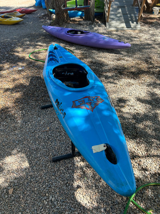 A blue Used Prijon Fly kayak from 4Corners Riversports with a planing hull sits on a metal stand over gravel, with a purple playboat and other kayaks visible nearby among trees.