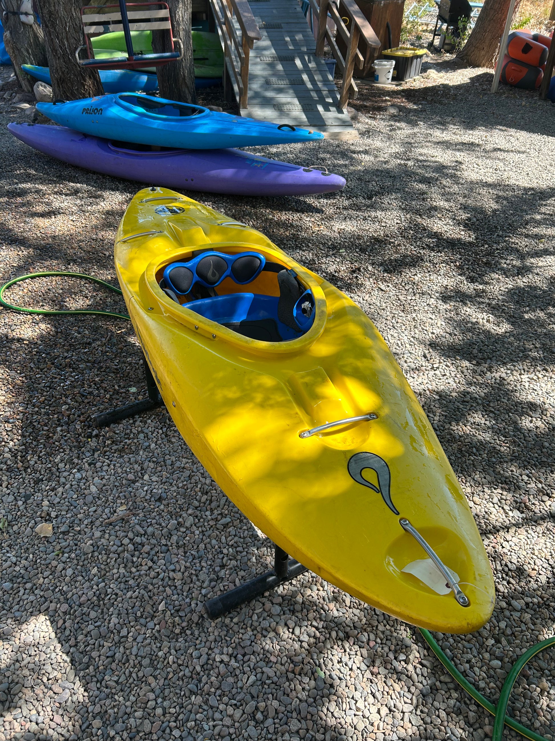 A Used LiquidLogic Trigger kayak from 4Corners Riversports in yellow, with black and blue paddles, sits on a stand over gravel; a purple playboat and wooden stairs are visible in the background.