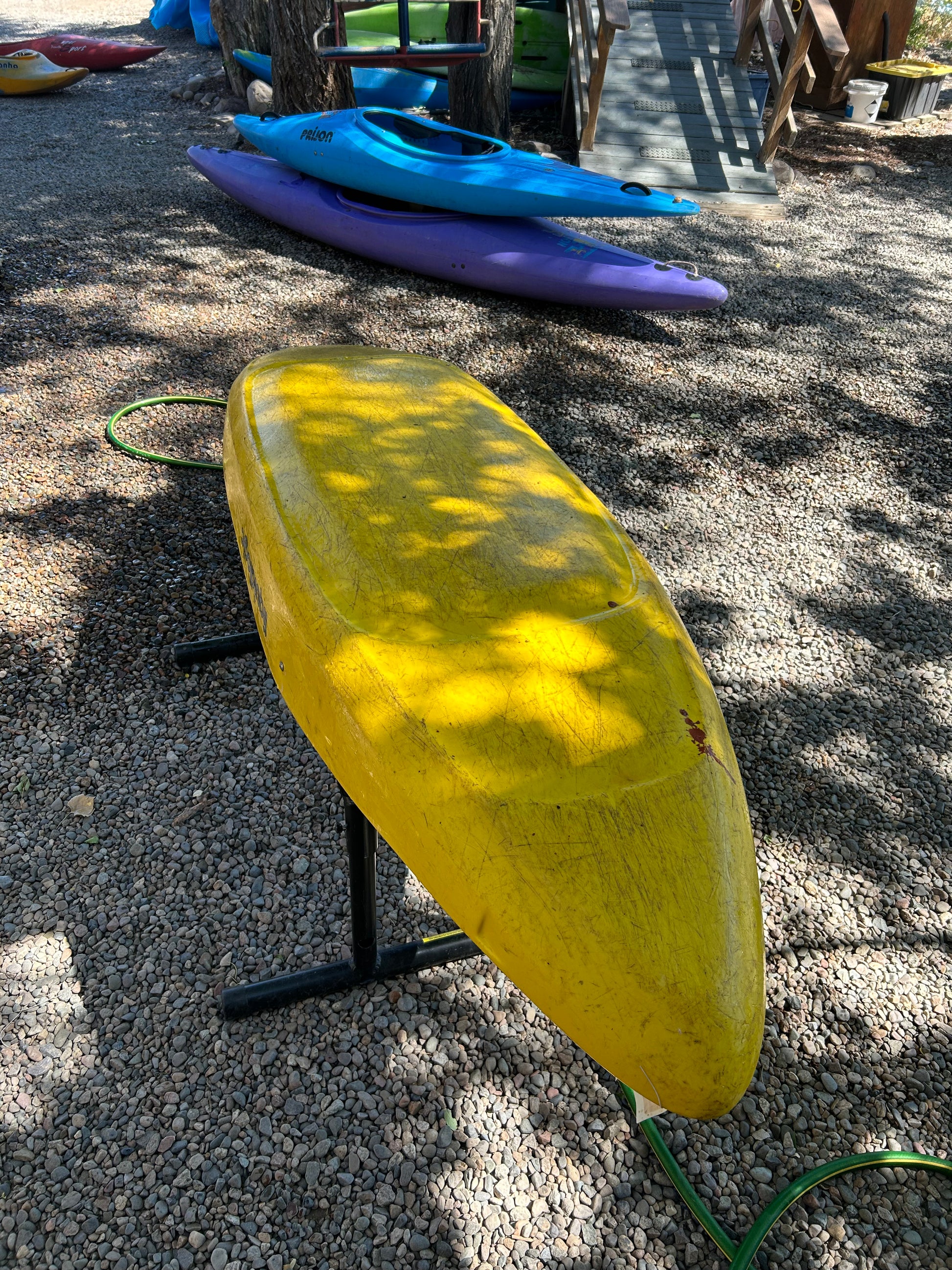 A yellow Used LiquidLogic Trigger from 4Corners Riversports rests upside down on a metal stand outdoors, with purple and blue kayaks nearby.