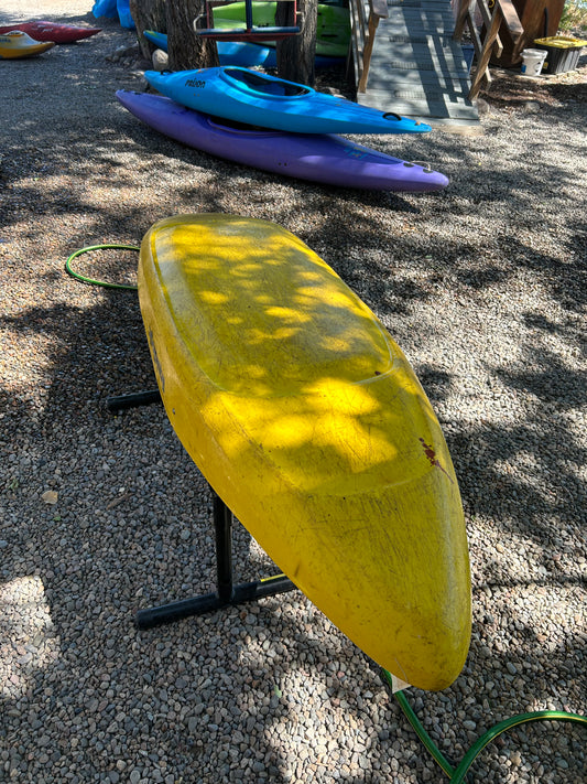 A yellow Used LiquidLogic Trigger from 4Corners Riversports rests upside down on a metal stand outdoors, with purple and blue kayaks nearby.